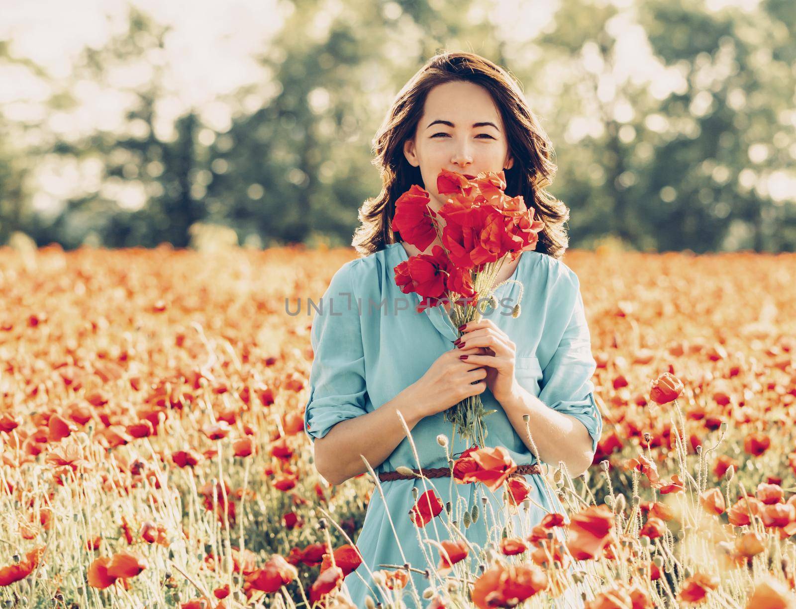 Beautiful young woman standing with bouquet of red poppies in flowers meadow in spring.