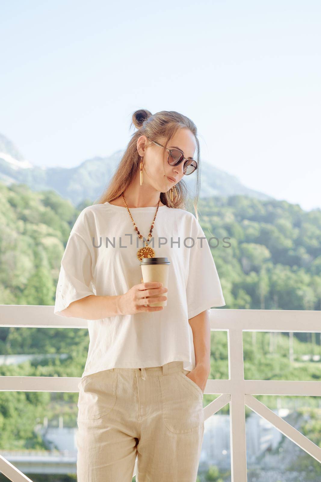 Stylish young woman wearing in white clothing with wooden accessories resting with cup of coffee on balcony in front of summer mountains.