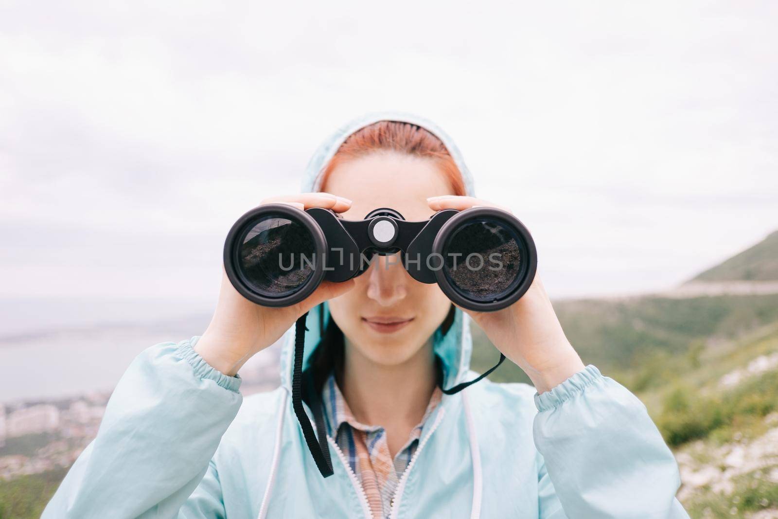Explorer hiker young woman looking through binoculars on background of summer mountains outdoor, front view. Travel and journey concept.