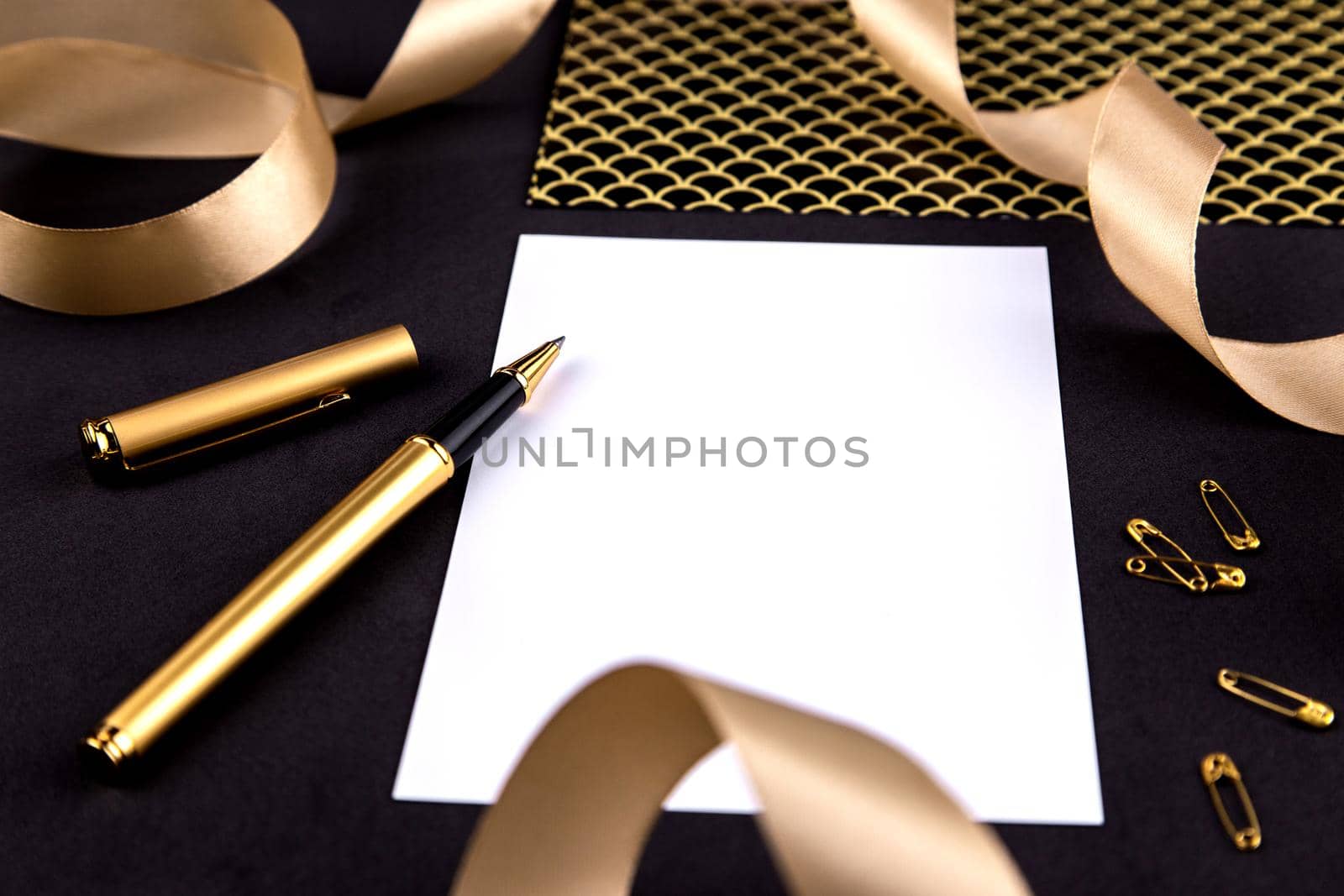 Gold pen, ribbon, paper clips and stationery on a black background with a white sheet of paper with copy space by lunarts