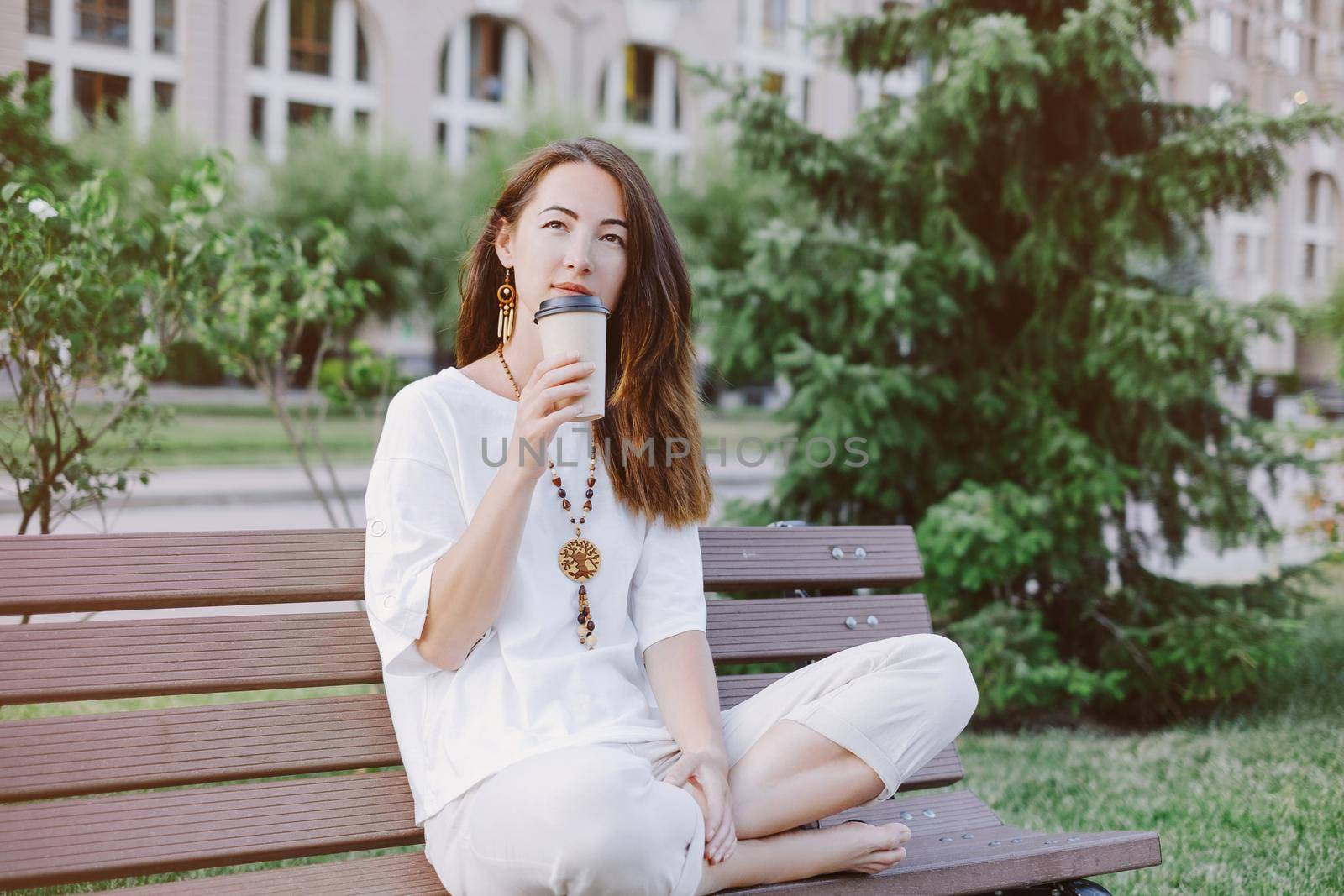 Beautiful young woman wearing in white clothing with wooden accessories sitting with paper cup of coffee and dreaming about something in summer park.