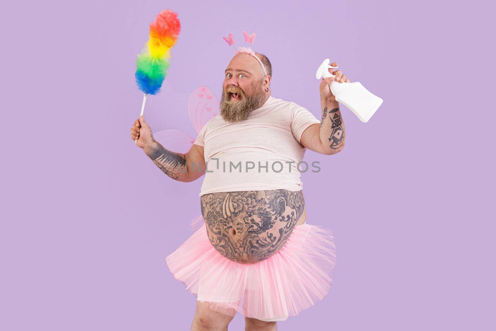 Man with overweight in fairy costume holds pp duster and spray bottle on purple background by Yaroslav_astakhov