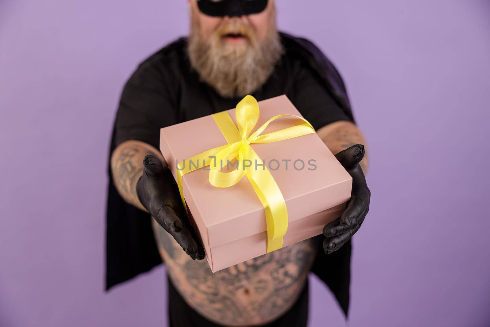Middle aged bearded man with overweight wearing carnval hero costume with cape on purple background in studio closeup, focus on hands with gift box
