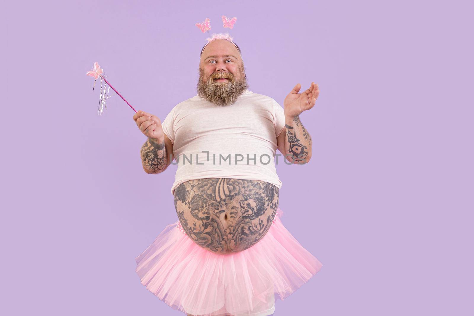 Smiling plus size mature man with tattooed abdomen wearing fairy suit holds magic stick with butterfly posing on purple background in studio