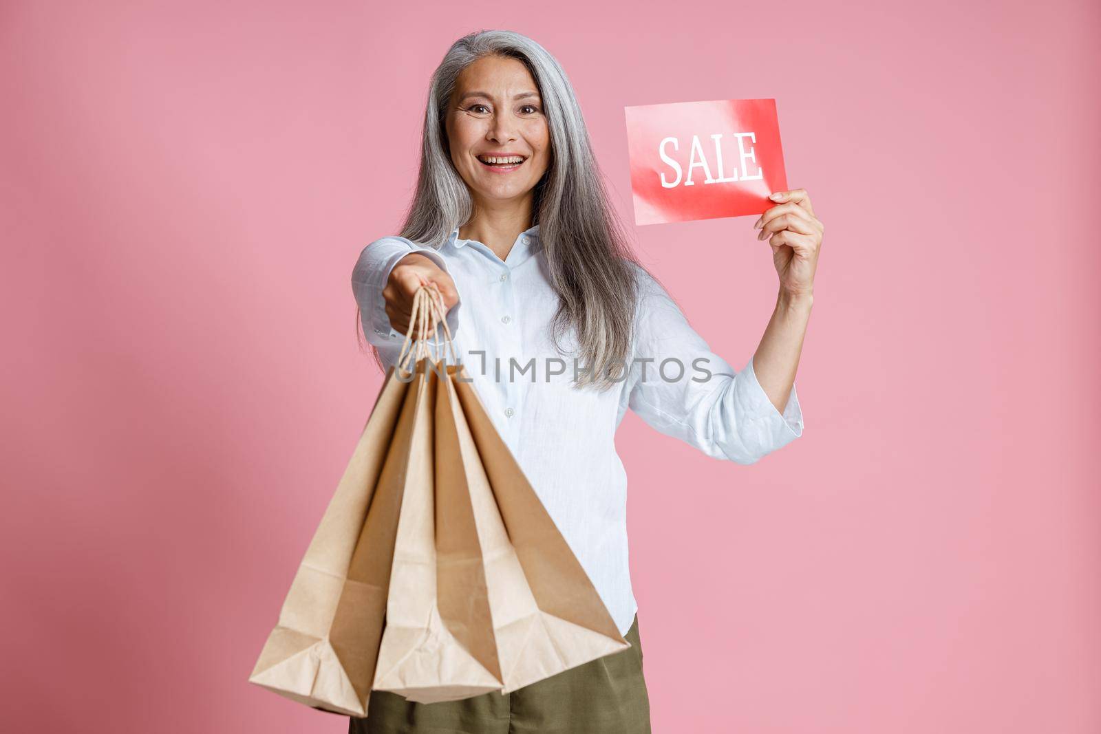 Pretty mature Asian woman holds paper shopping bags and red card with word Sale posing on pink background in studio