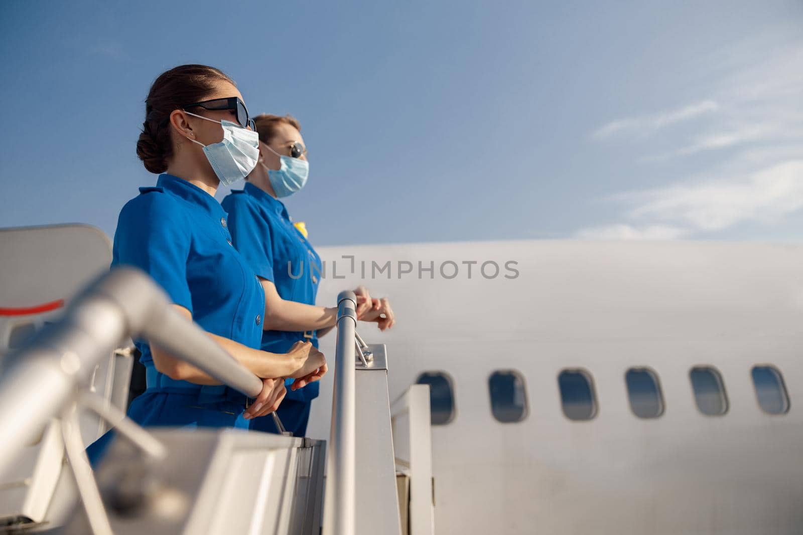 Side view of two young air stewardesses in blue uniform, sunglasses and protective face masks looking away, standing on airstair during boarding. Aircrew, occupation, covid19 concept