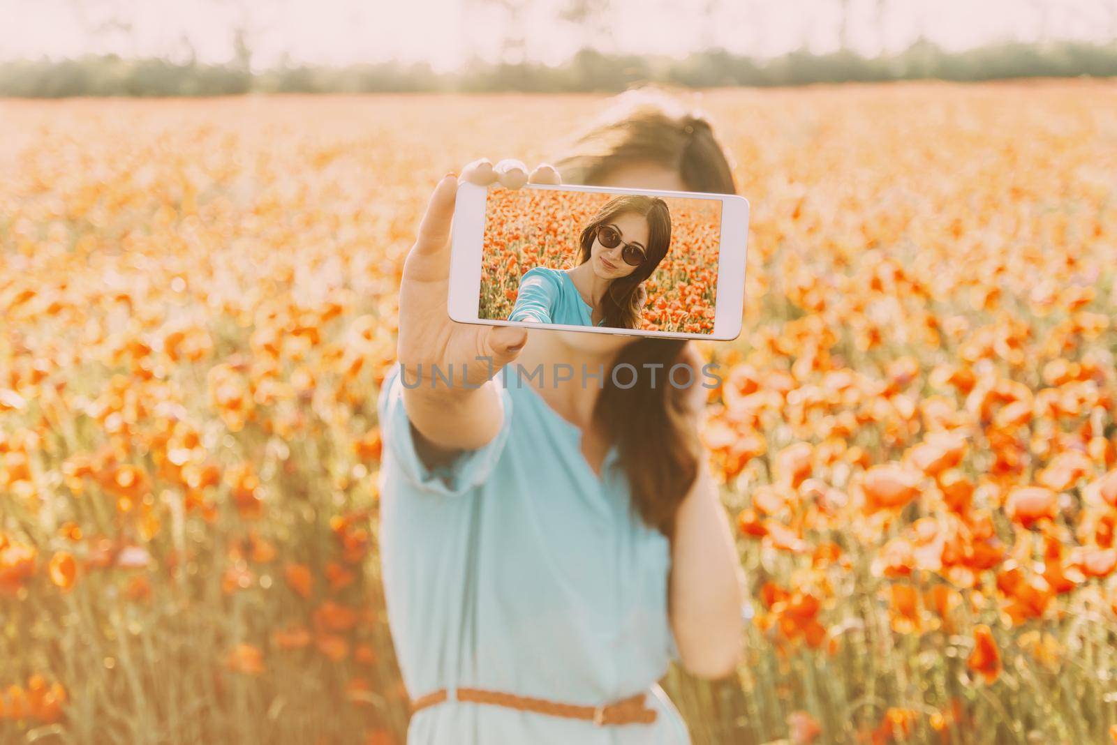 Beautiful brunette young woman in glasses taking a photo selfie with smartphone in flowers meadow in summer outdoor.