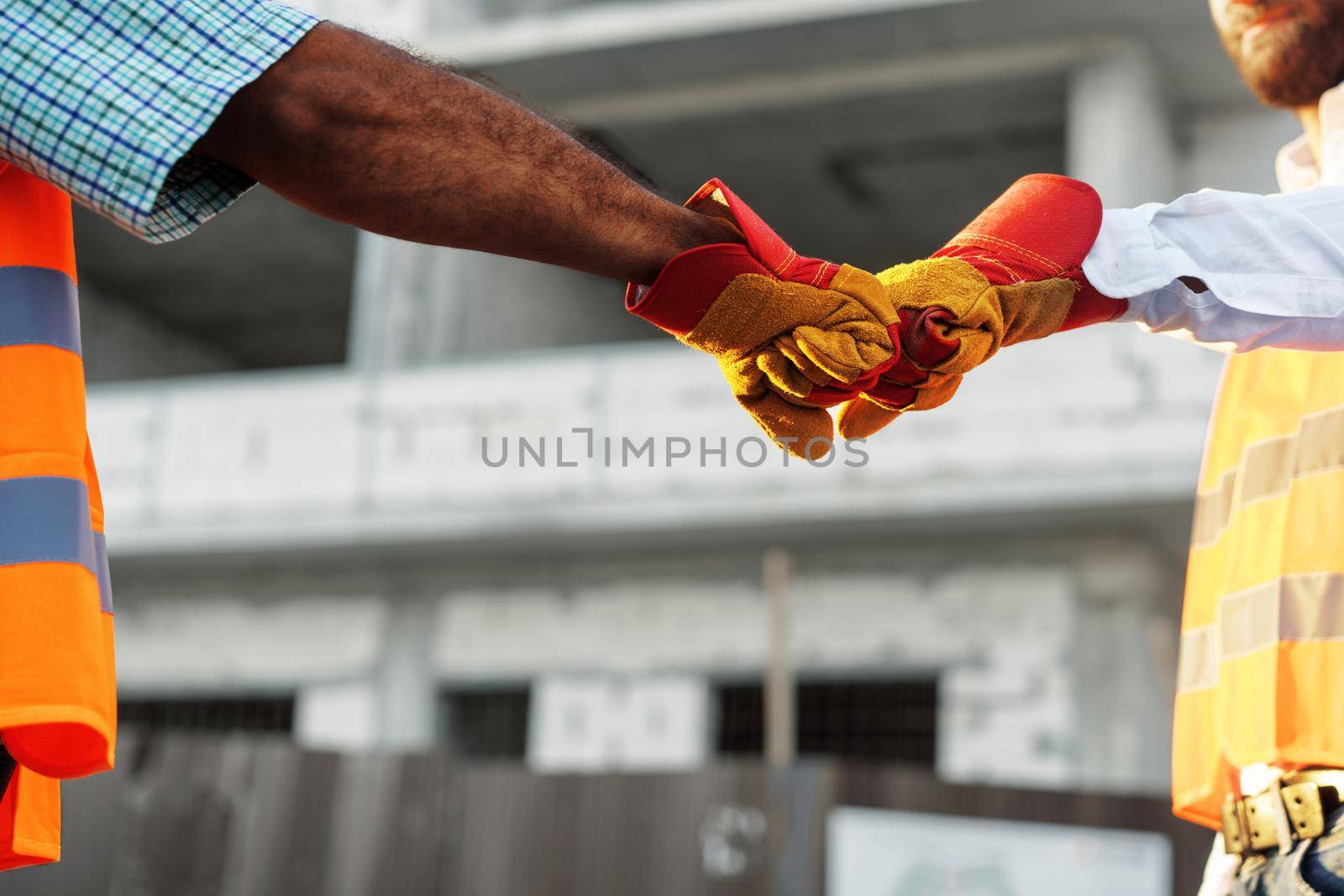 Two men engineers in workwear shaking hands against construction site, close up