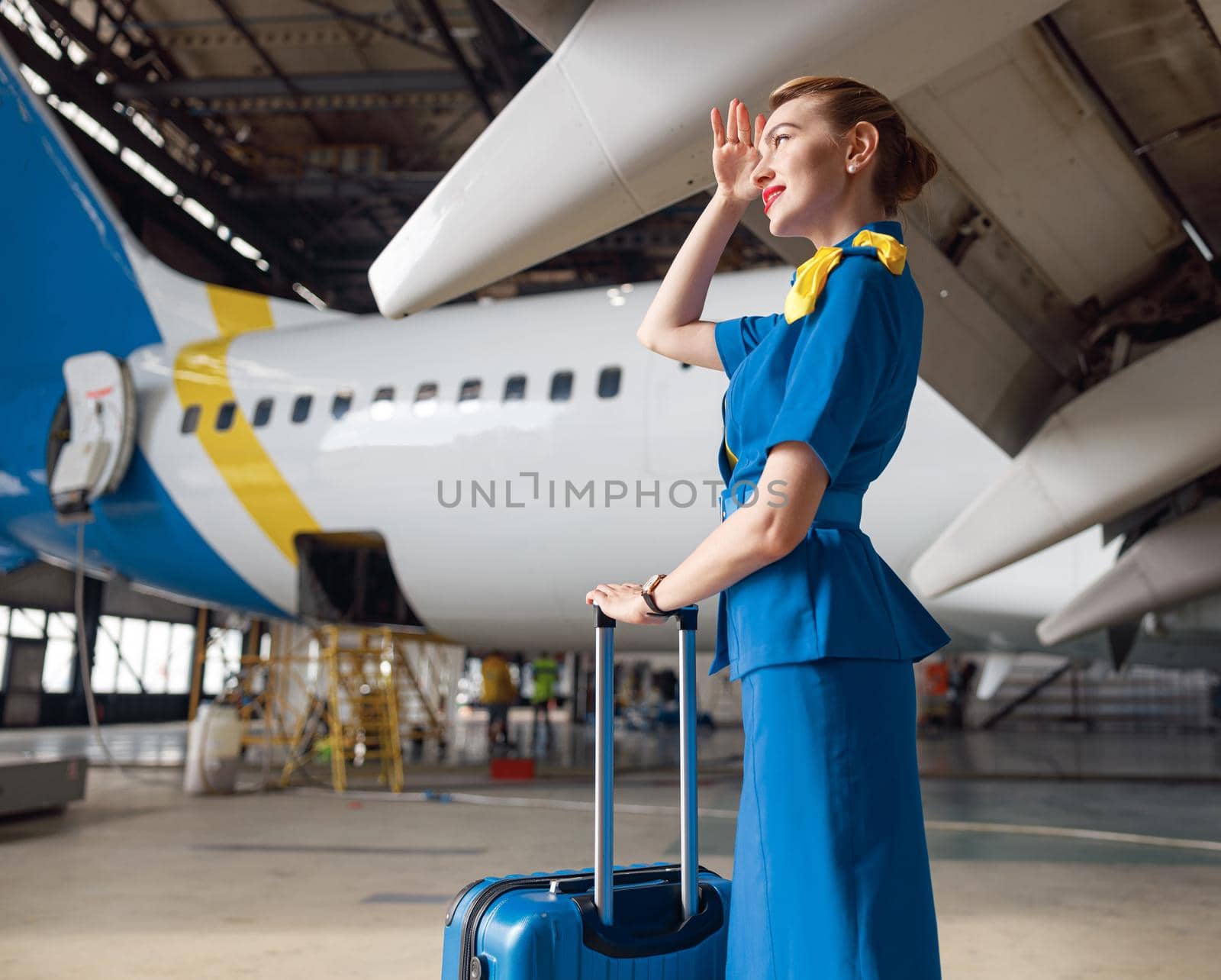 Beautiful air stewardesses in bright blue uniform looking away while standing with suitcase in front of passenger aircraft by Yaroslav_astakhov