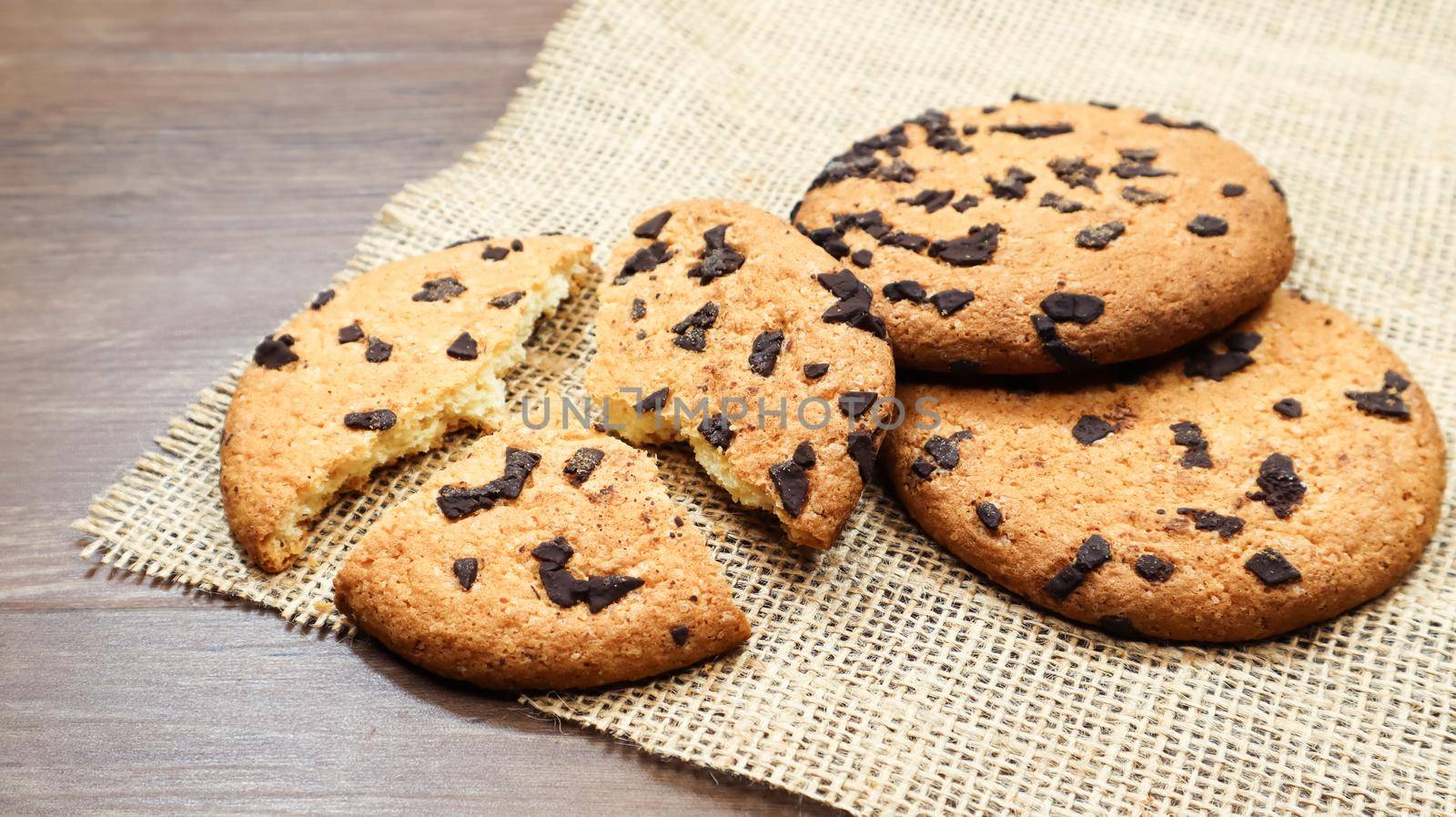 American chocolate chip cookies on a brown wooden table and on a linen napkin close-up. Traditional rounded crunchy dough with chocolate chips. Bakery. Delicious dessert, pastries. Rural still life