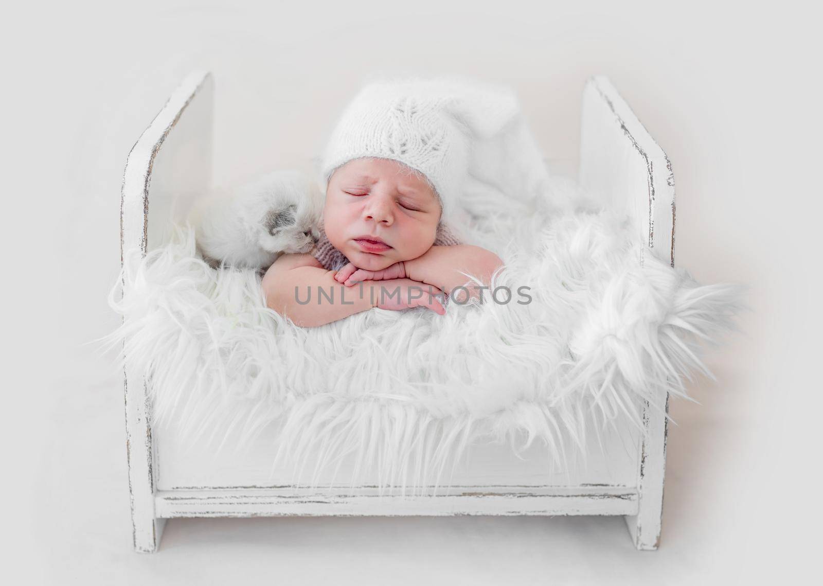 Adorable newborn baby boy sleeping on his tummy in white wooden bed with fur with little fluffy kitten. Cute infant kid wearing knitted hat napping with cat kity during studio photoshoot