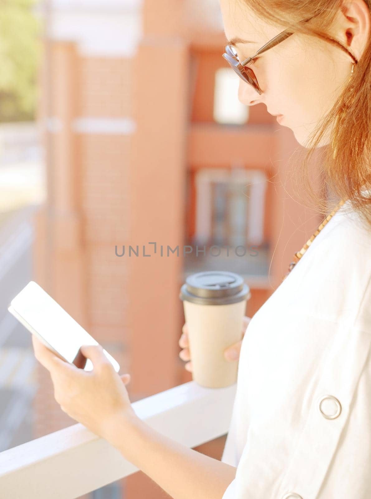 Young woman using smartphone while resting with cup of coffee on balcony in summer morning, city lifestyle.