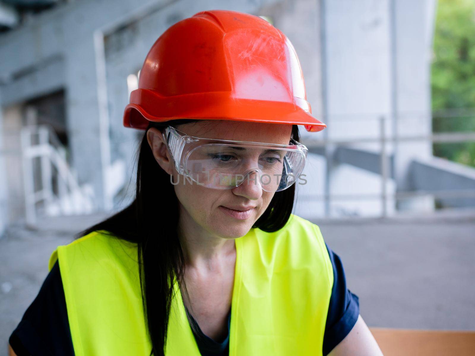 Woman architect in protective helmet and goggles working on personal computer on new project at construction site by Utlanov