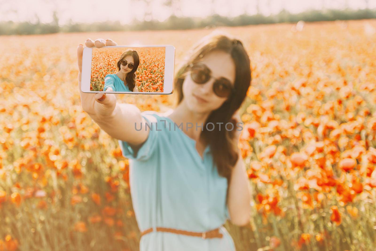 Beautiful smiling young woman in glasses taking a photo selfie with smartphone in flowers meadow in summer outdoor.