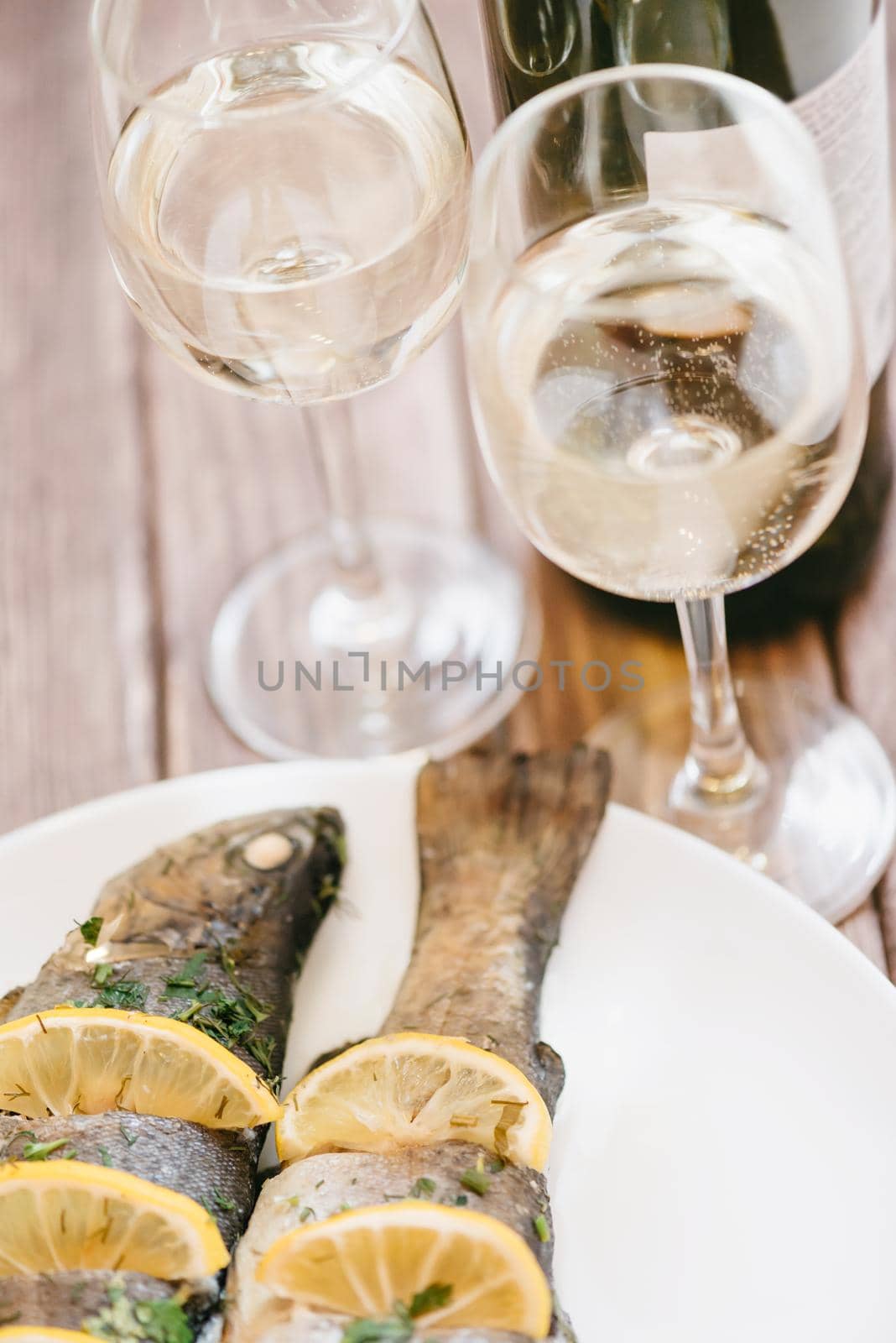 Baked fish trout dish with lemons and bottle with two glasses of white wine for couple on wooden table.
