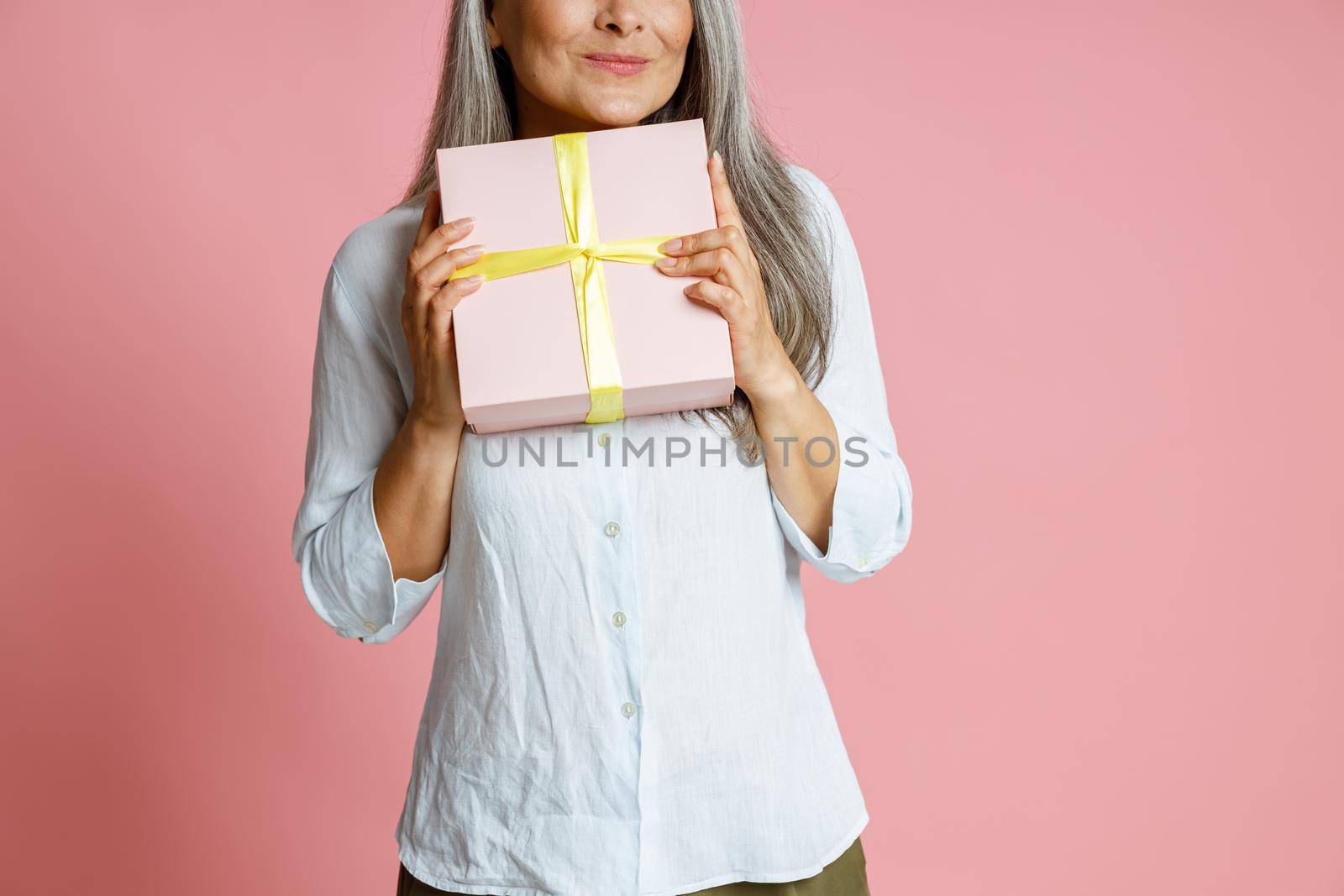 Positive mature female model with long silver hair holds gift box posing on pink background in studio closeup