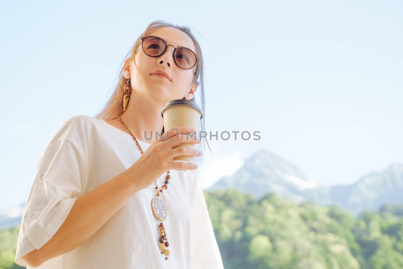 Beautiful stylish young woman holding cup of coffee in morning on background of summer mountains.