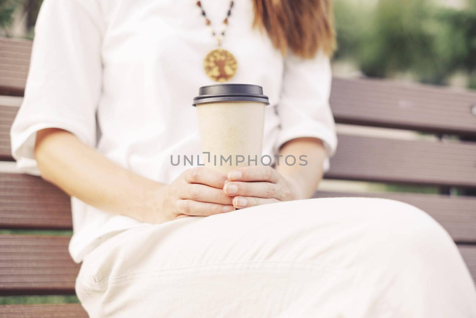 Unrecognizable woman wearing in white clothing sitting with takeaway paper cup of coffee on wooden bench.