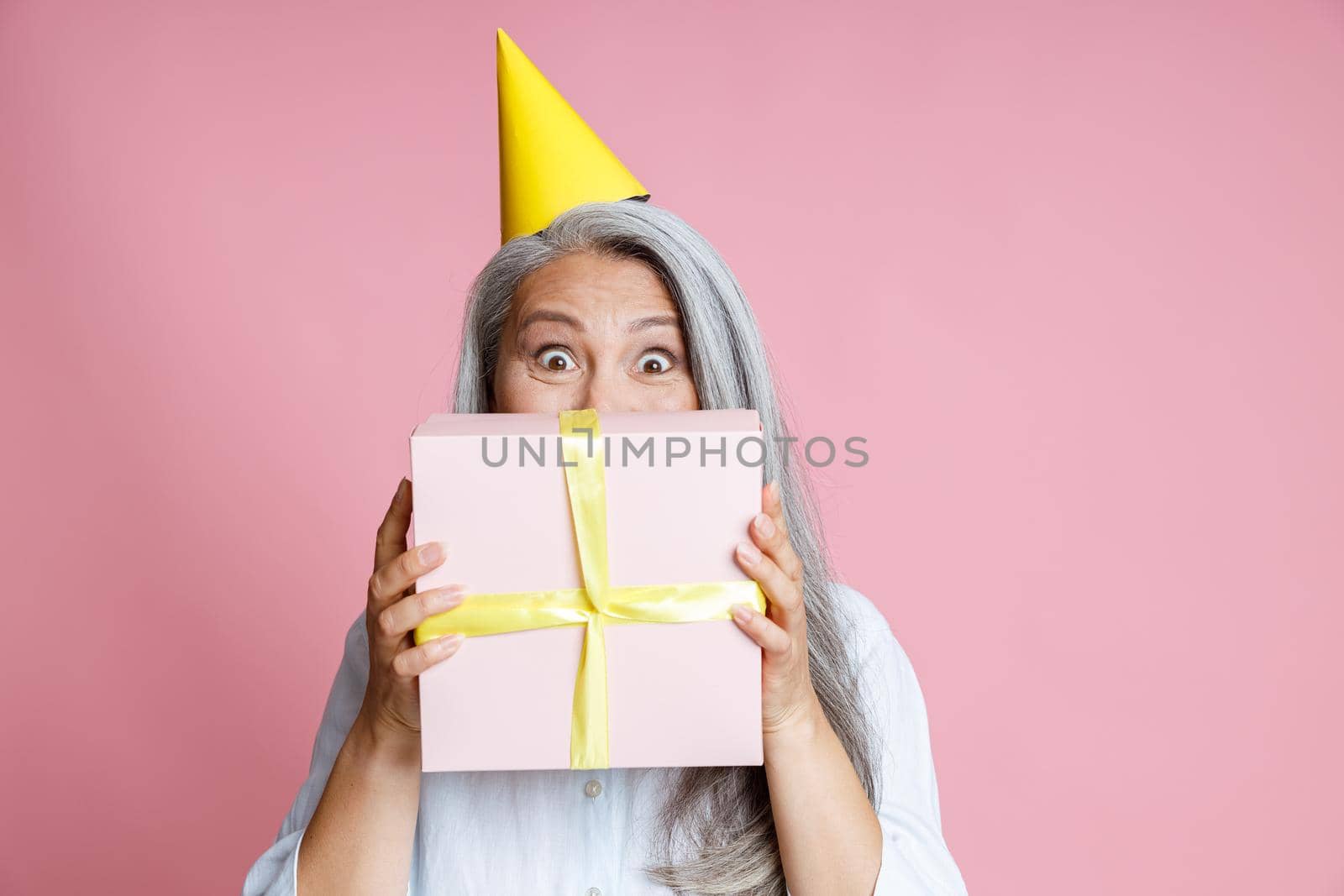 Happy middle aged Asian woman with grey hair and yellow party hat holds gift box near face on pink background in studio