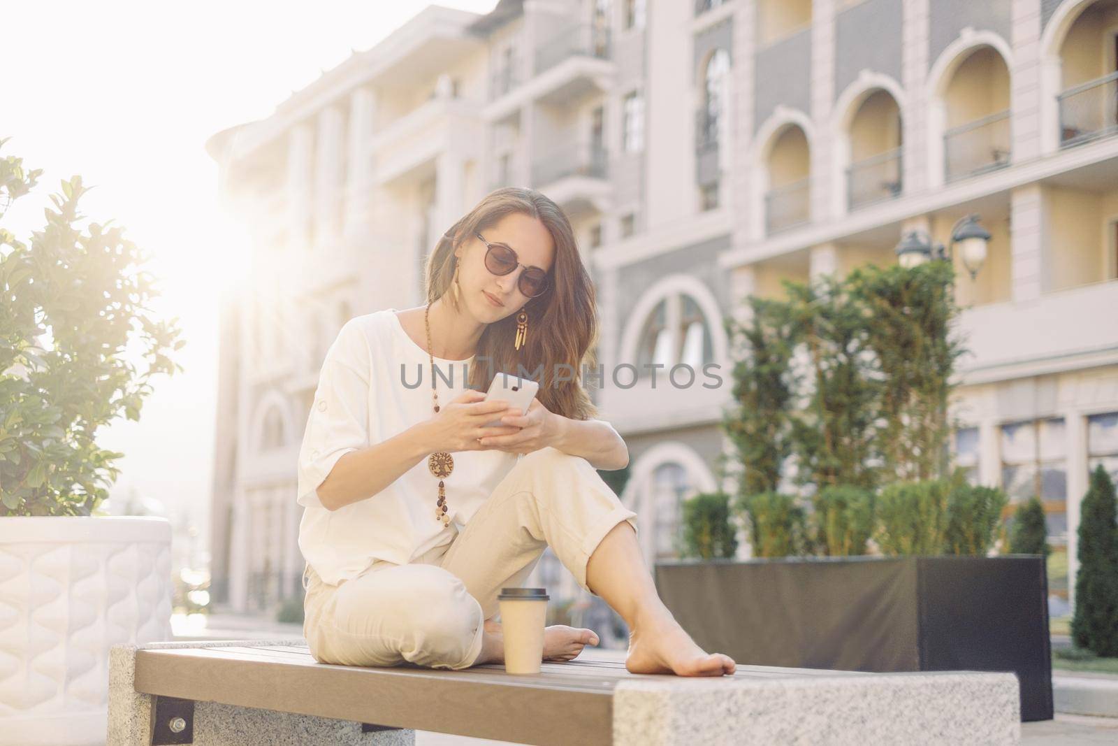 Smiling beautiful young woman using smartphone while resting on wooden bench in city street in summer outdoor.