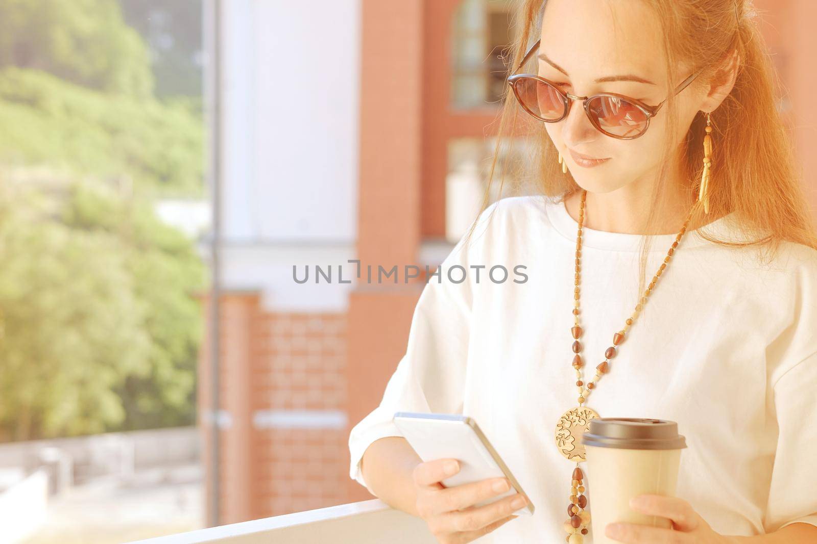 Beautiful boho stylish young woman using mobile phone while drinking coffee on balcony in spring morning, city lifestyle.