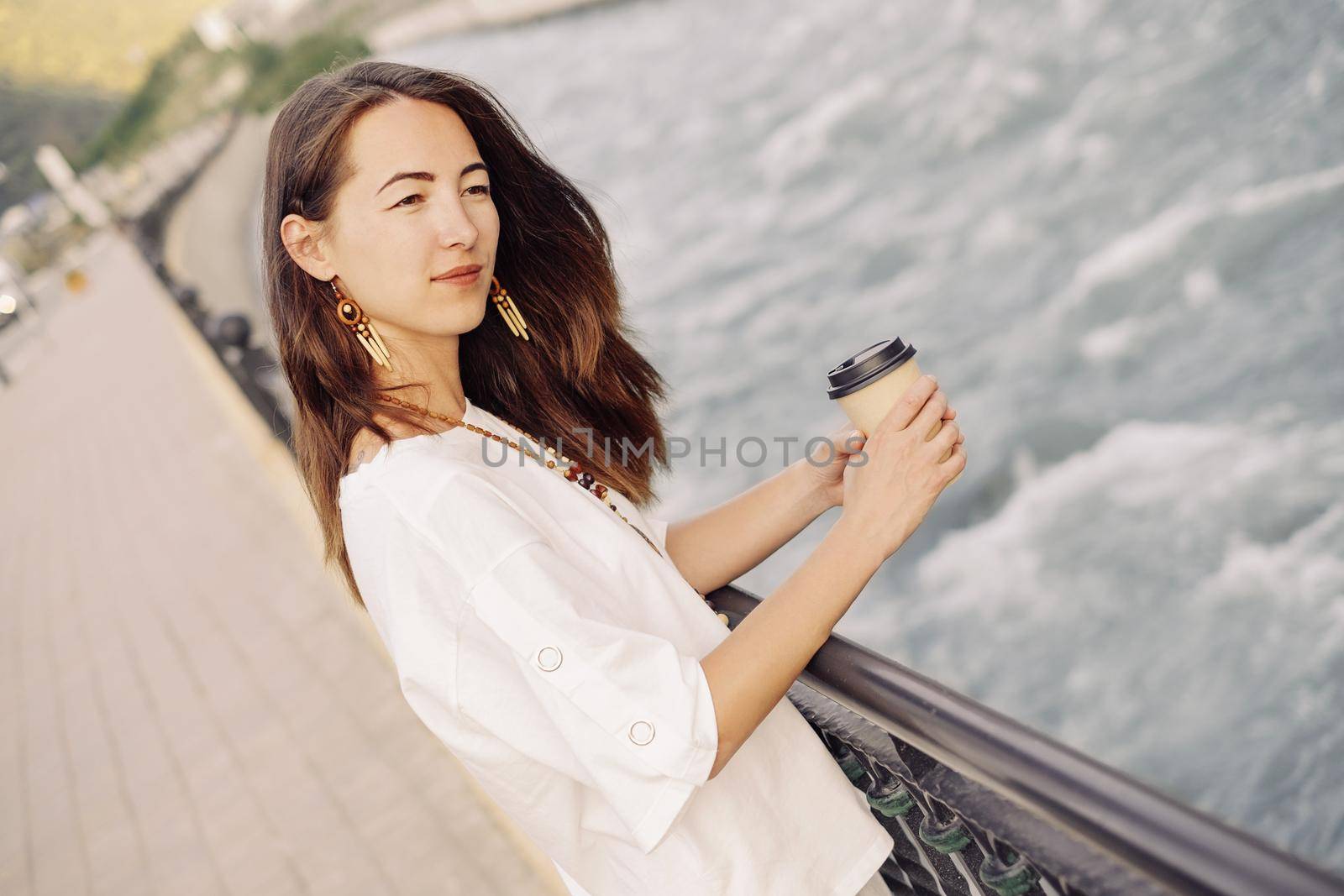 Smiling beautiful young woman walking with takeaway paper cup of coffee on embankment of river in summer outdoor.