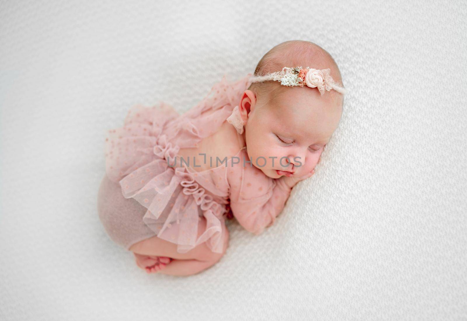 Newborn baby girl wearing cute costume sleeping and holding her hands under cheek. Little cute infant child napping lying on her tummy on white fabric. Adorable kid during studio photoshoot