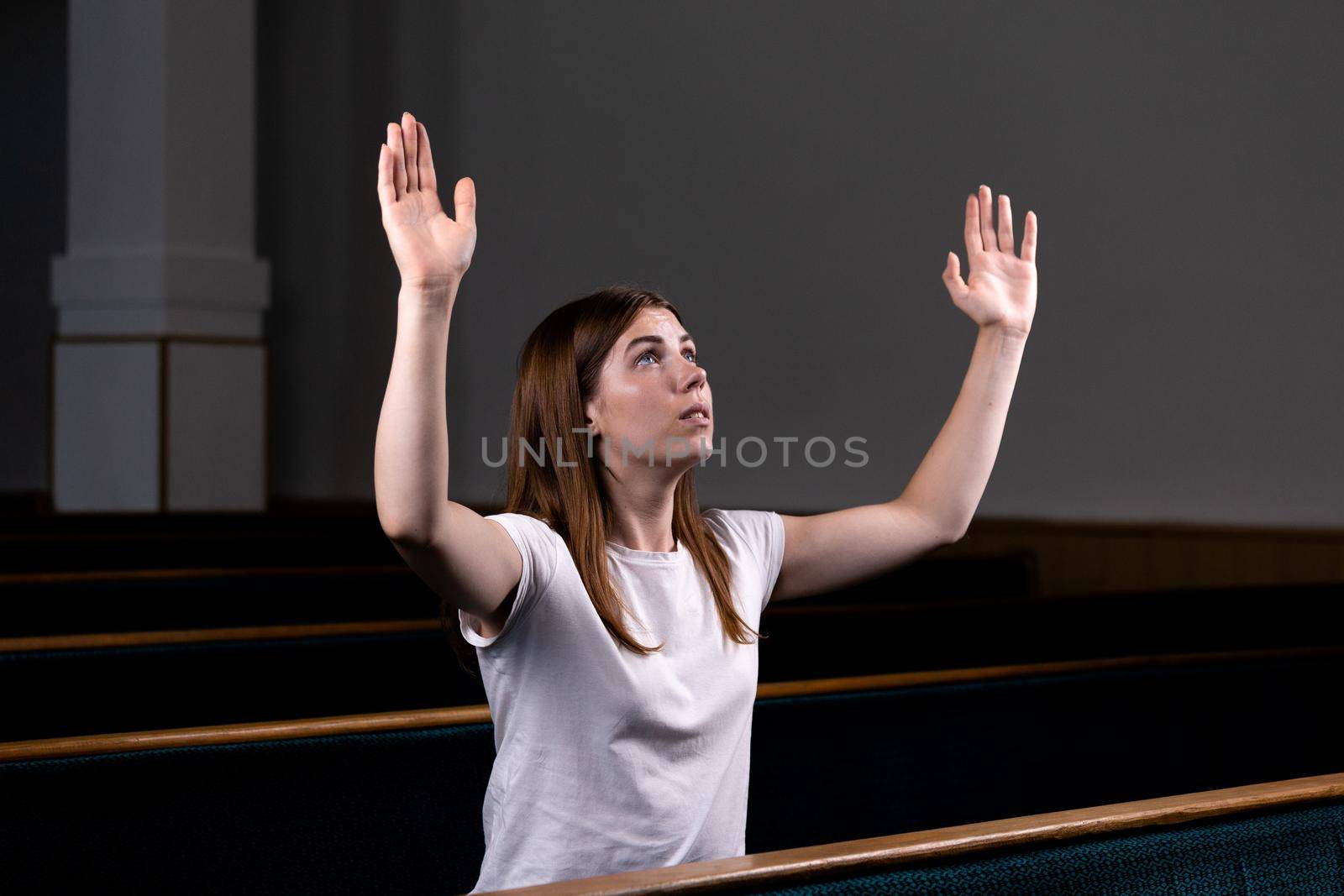 A Christian girl in white shirt is sits with his hands up and face and praying with humble heart in the church by lunarts