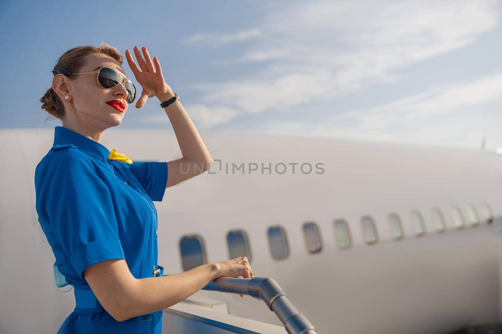 Portrait of elegant air stewardess in blue uniform and sunglasses holding hand near her head and looking far away, standing on airstair on a daytime by Yaroslav_astakhov