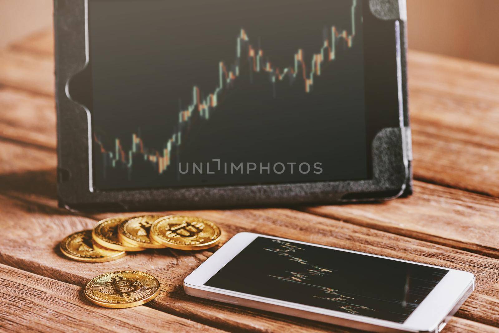 Bitcoins cryptocurrency coins on wooden table near mobile phone and digital tablet with growth chart on screen.