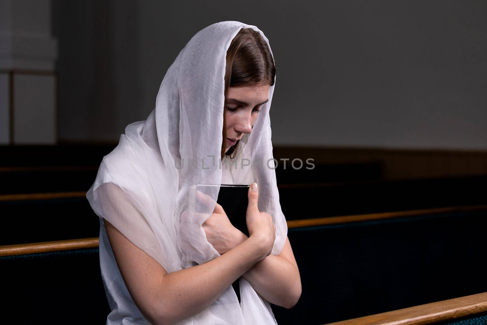 A young modest girl with a handkerchief on her head and a bible in her hands is sitting in church and praying. The concept of religion, prayer, worship by lunarts