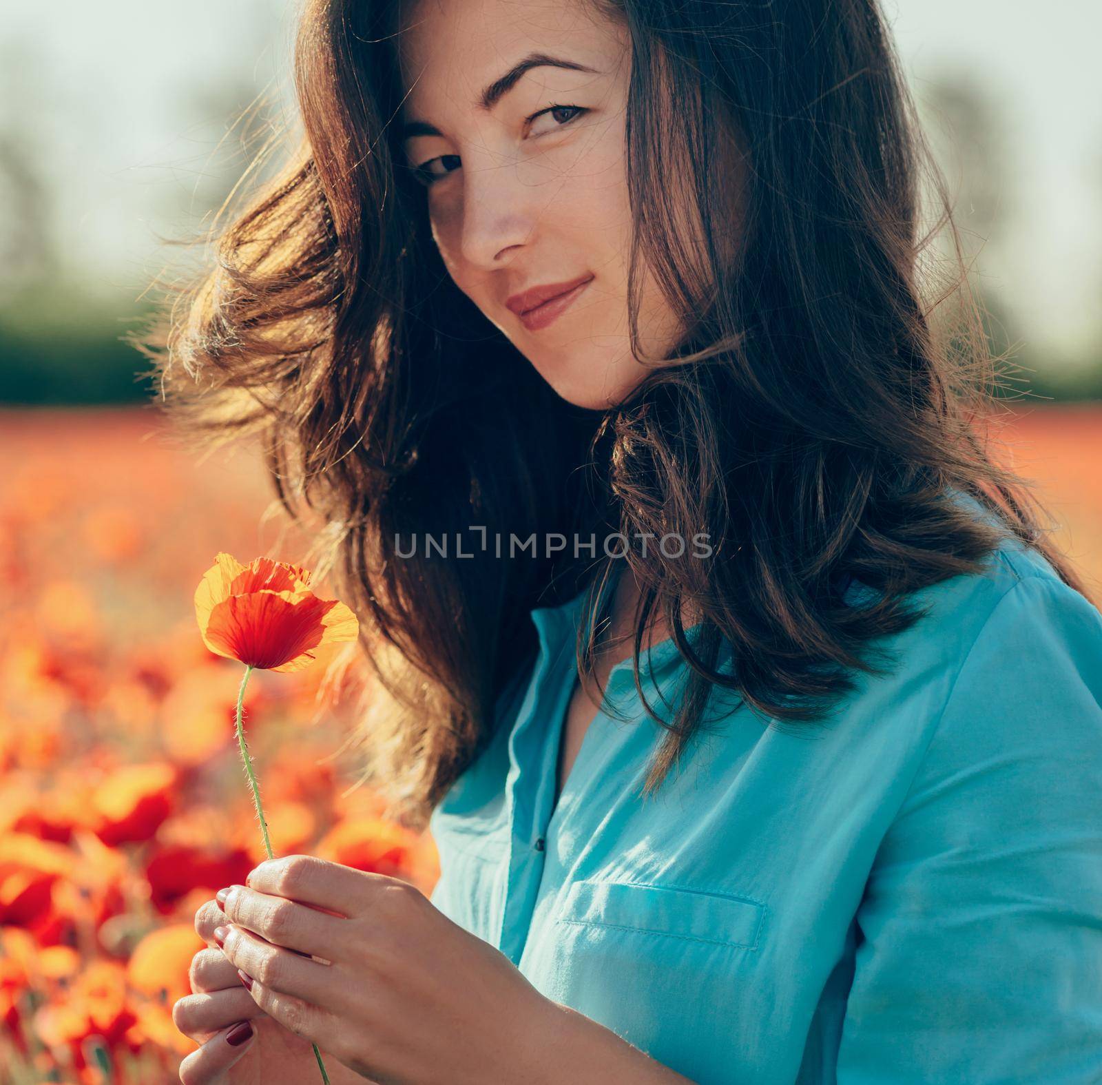 Outdoor portrait of smiling attractive brunette young woman with red poppy flower in flowers meadow, looking at camera.