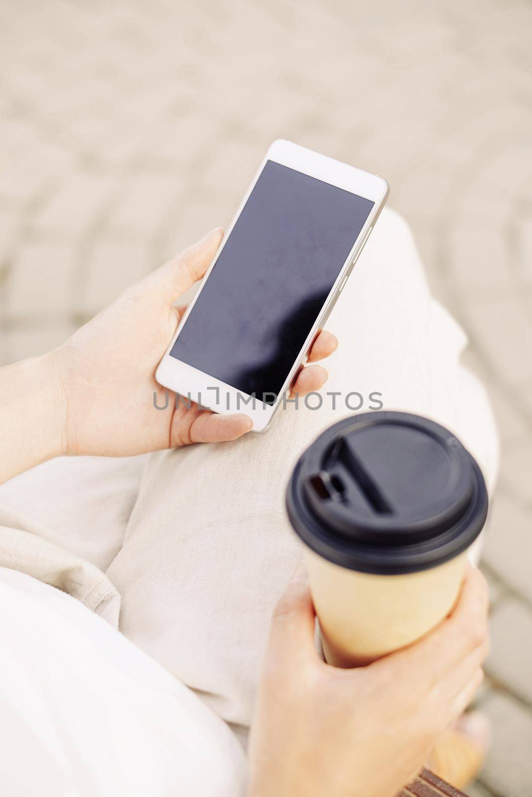 Unrecognizable young woman sitting with takeaway paper cup of coffee and smartphone, mock-up.