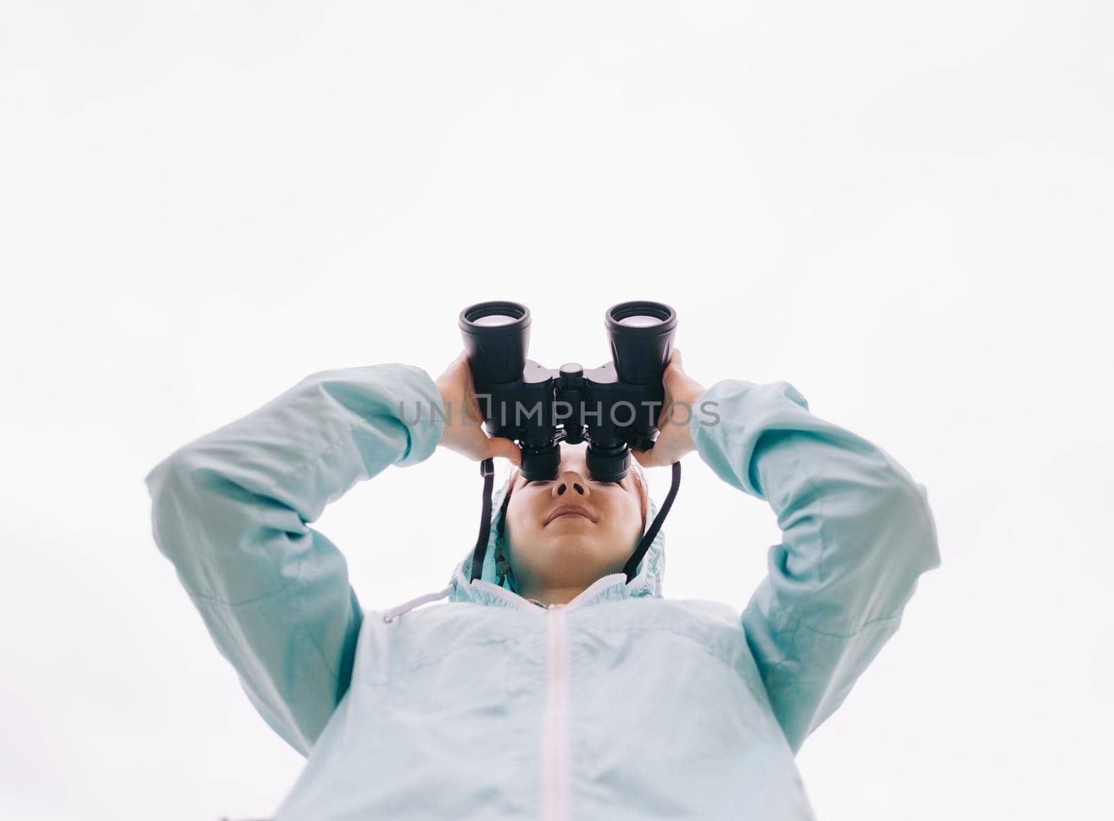 Bottom view of traveler young woman watching with binoculars and exploring surroundings on white background of sky.
