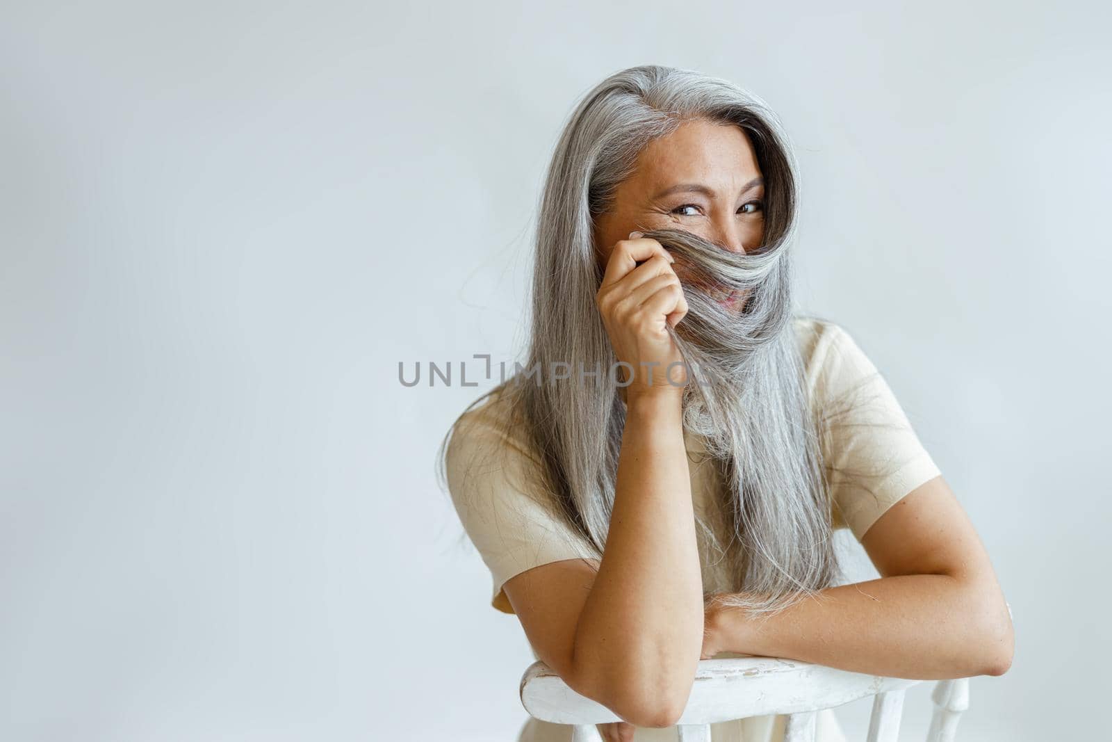 Positive middle aged Asian lady uses hoary hair as niqab sitting on chair on light background in studio, space for text. Mature beauty lifestyle