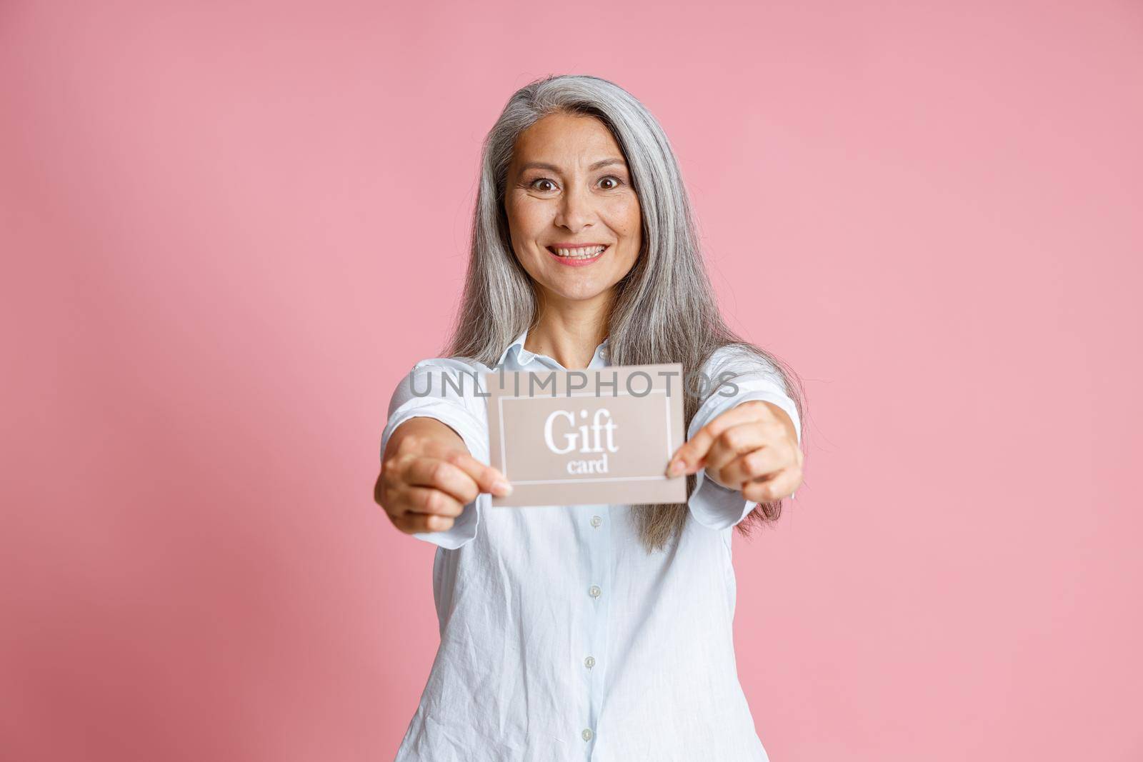 Attractive grey haired Asian lady holds gift card on pink background by Yaroslav_astakhov