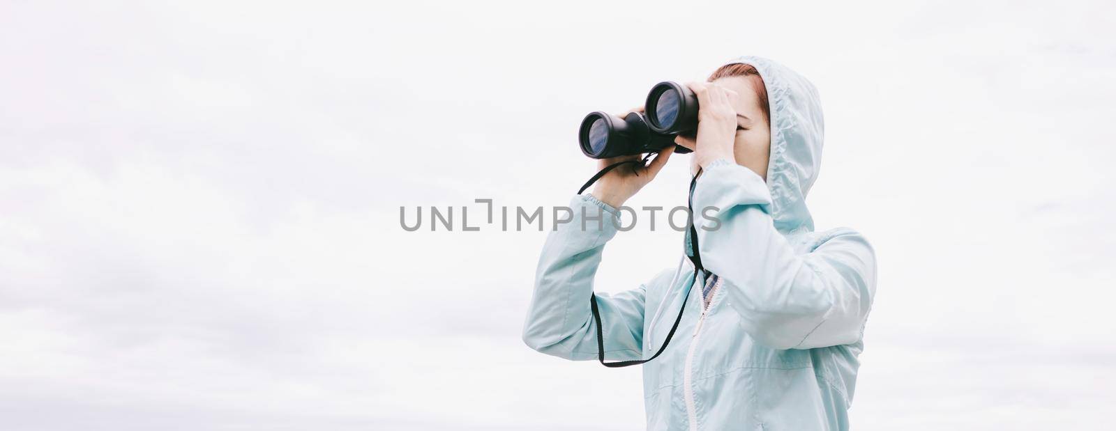 Traveler young woman looking through binoculars on background of sky. Copy-space in left part of image.
