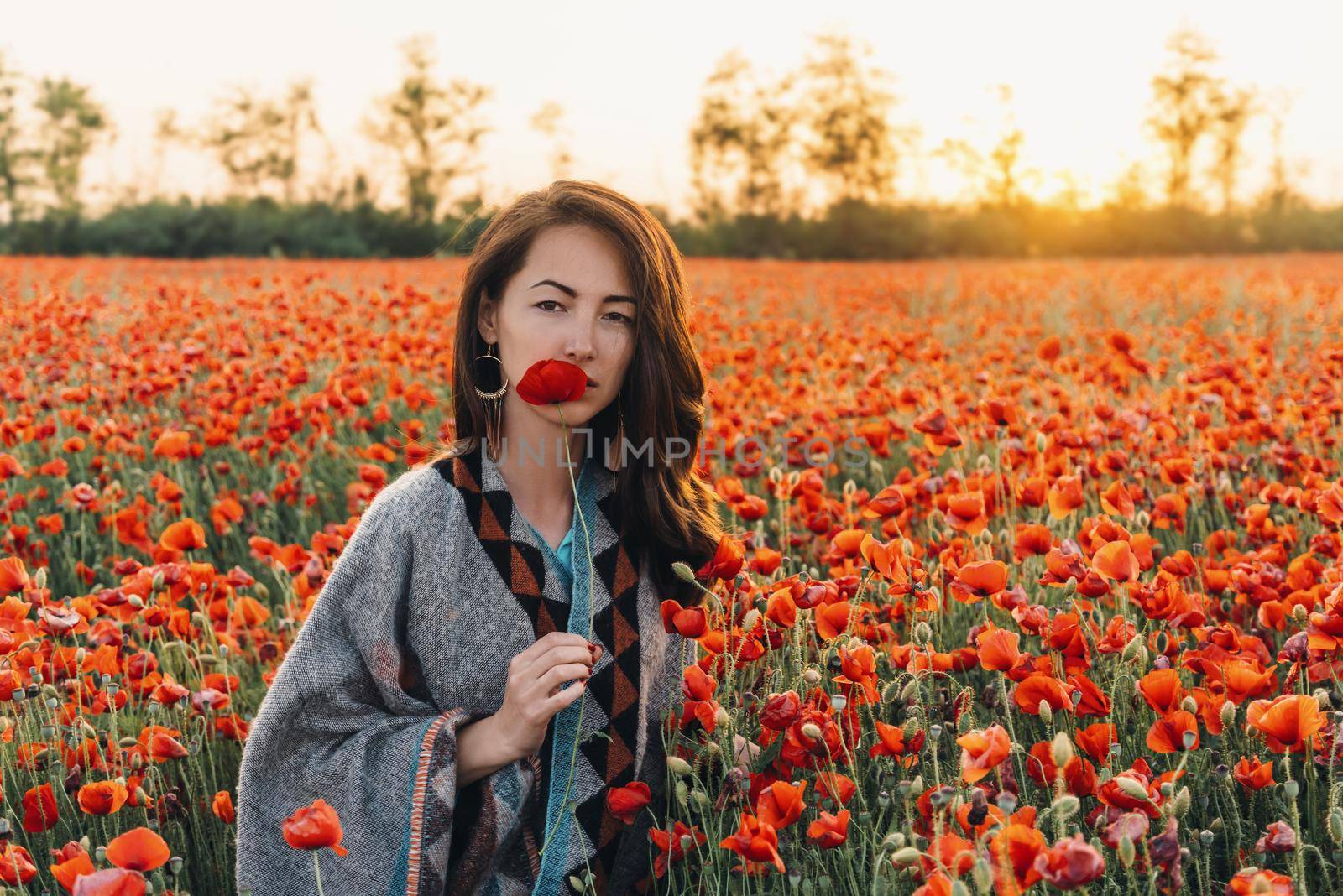 Fashion outdoor portrait of pretty brunette young woman holding red poppy near her face in flower meadow at sunset, looking at camera.