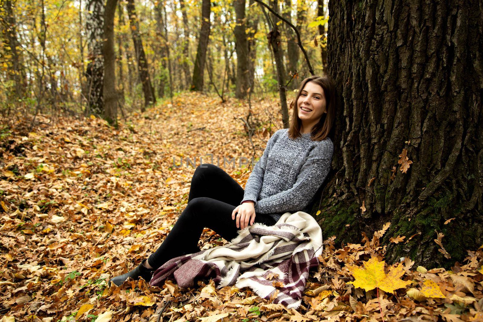 Cute girl in a gray jacket sitting in the autumn forest on a yellow leaf near a large tree by lunarts