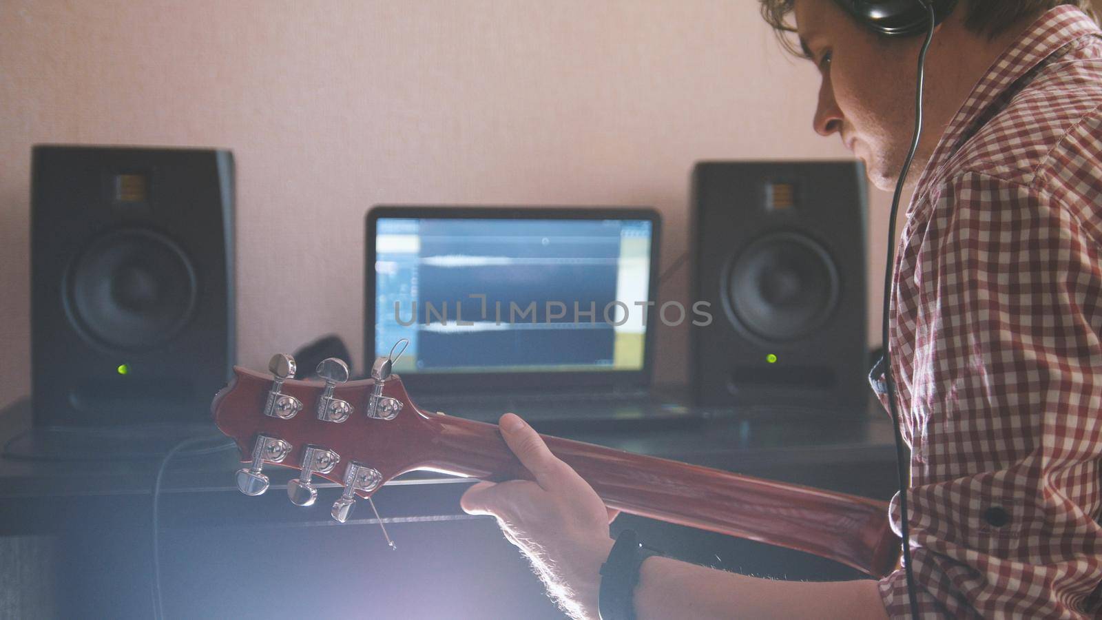 Young musician composes and records music playing the electric guitar, using computer and keyboard, from the front view