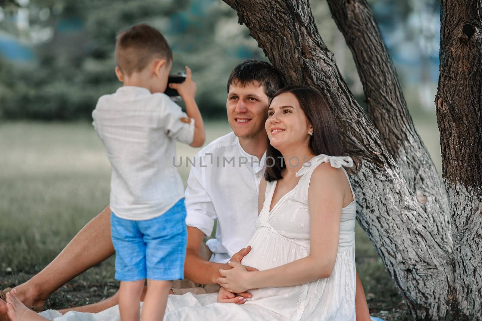 A happy family is resting under a tree on a summer day. The boy takes pictures of his mother and father, who are cheerful and smiling.