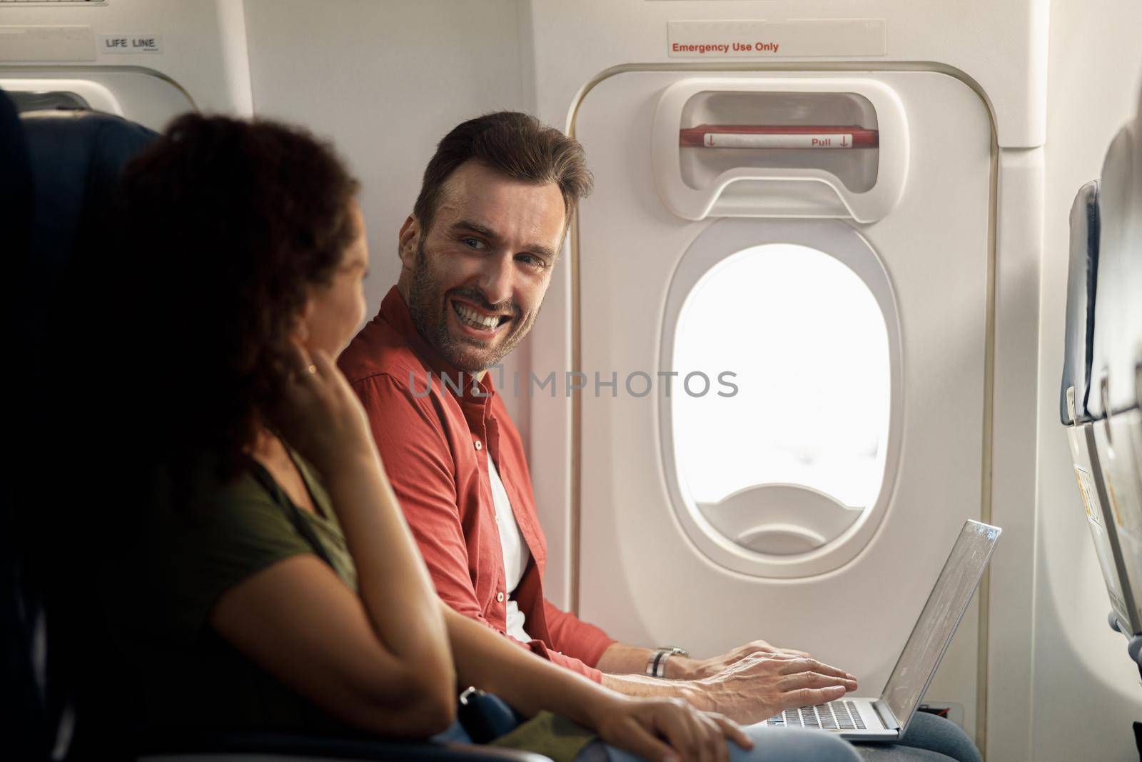 Cheerful caucasian man smiling to his wife while using laptop computer, sitting on the airplane by Yaroslav_astakhov