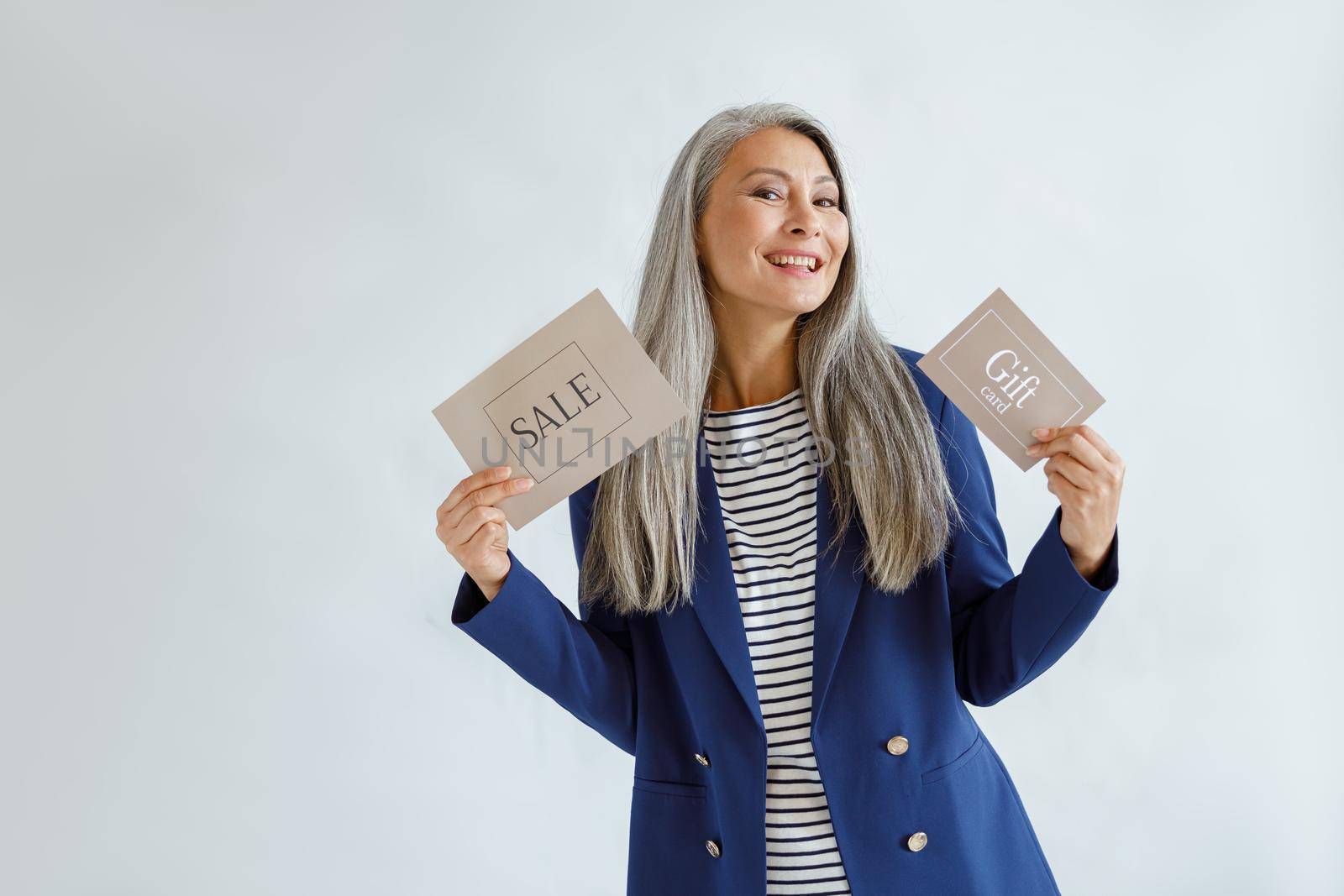 Cheerful middle aged Asian woman with hoary hair holds gift card and Sale sign standing on light grey background in studio, space for text