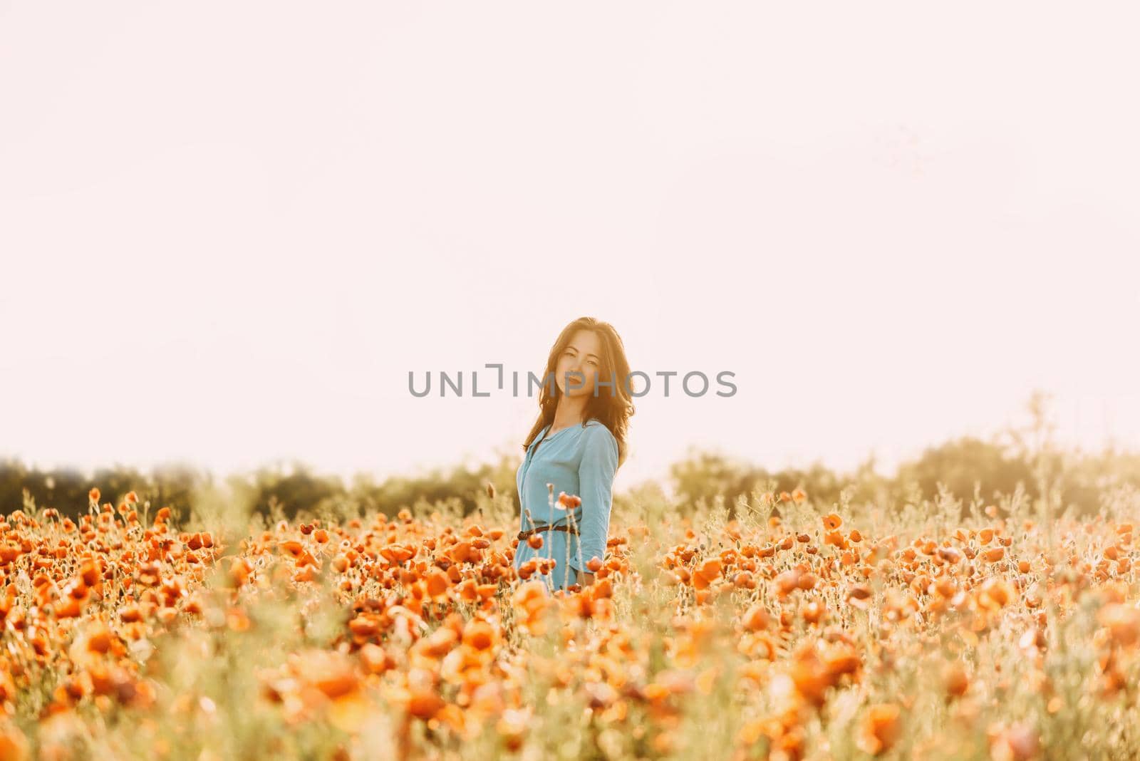 Beautiful brunette young woman with long hair walking in red poppy flower field on sunny summer day.