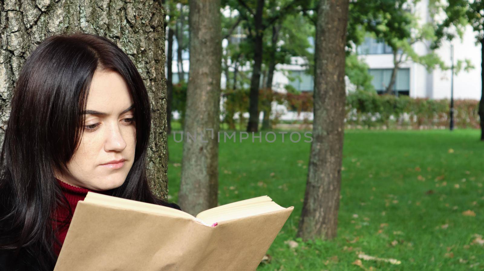 Portrait of a beautiful young woman who is sitting under a tree and reading her favorite book in a city park on green grass on a pleasant sunny day. Vacation, education and study concept