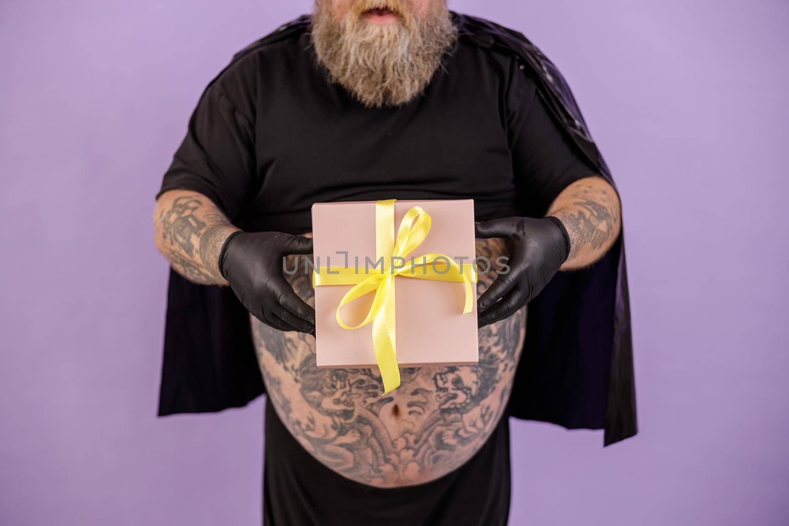Mature bearded plus size man wearing hero suit with cape on purple background in studio closeup, focus on hands with gift box