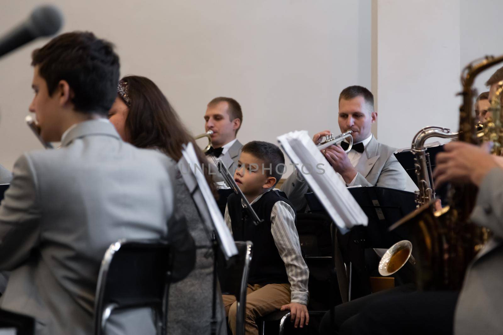 KOROSTEN - NOV, 10, 2019: A little boy sits in an orchestra near the musicians play different musical instruments in gray suits.