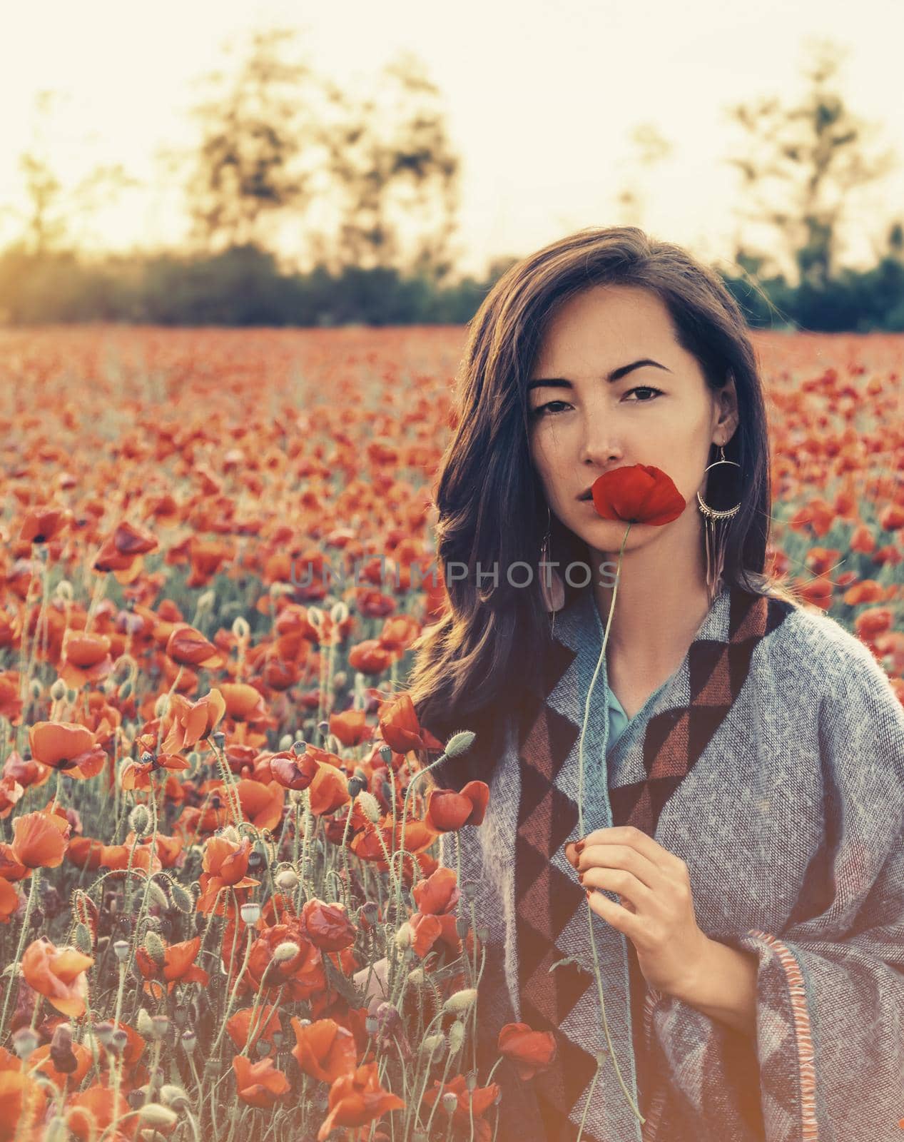 Beauty outdoor portrait of brunette young woman with red poppy near her face in flower meadow, looking at camera.