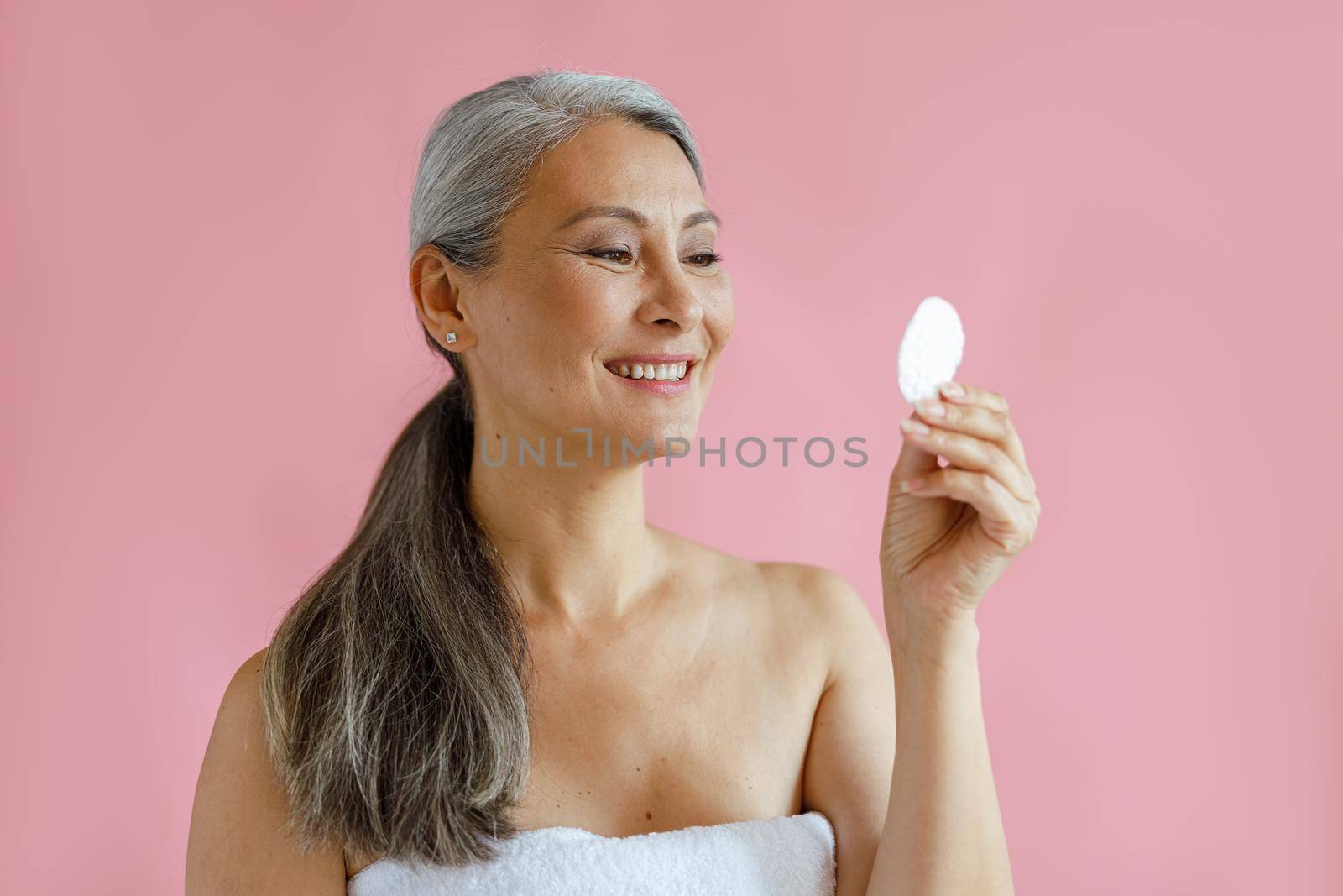 Beautiful silver haired Asian woman looks at cotton pad standing on pink background in studio. Mature beauty lifestyle