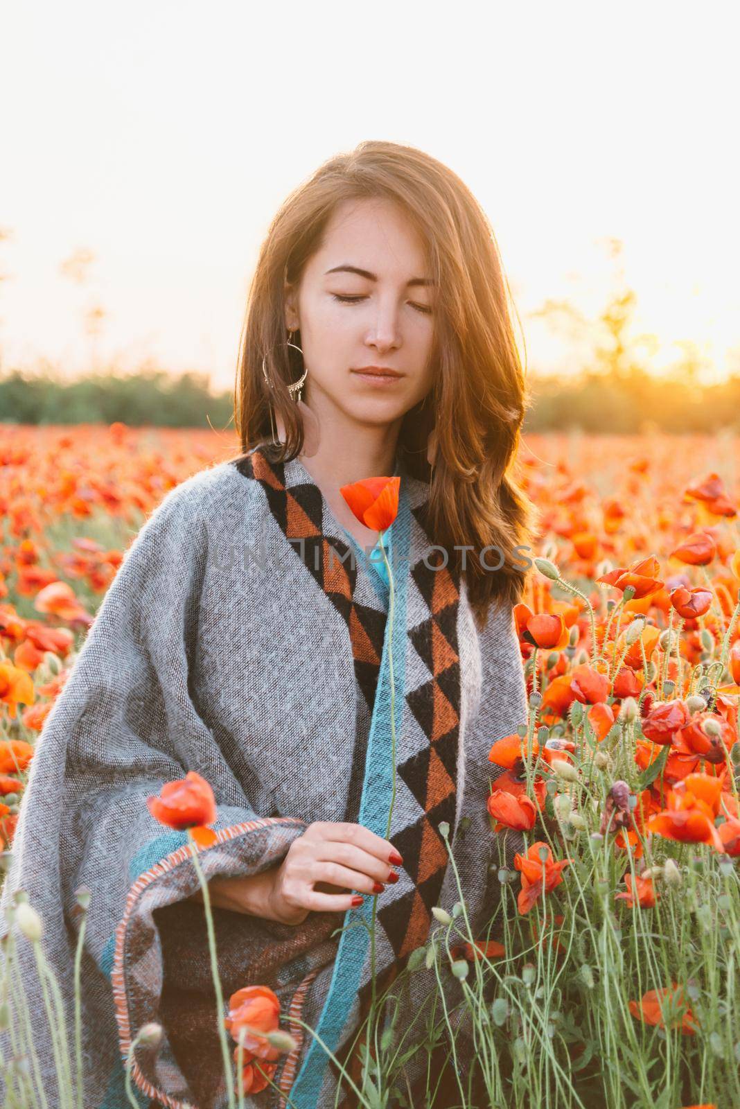 Beautiful brunette young woman with closed eyes relaxing in red poppy flower field at sunset in summer.
